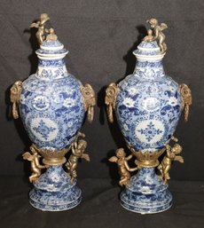 Pair Of Blue And White Glazed Porcelain Urns With Floral Design, Bronze Cherubs On Lids And Bases, And Rams He